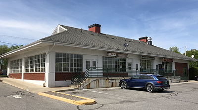 Single-story brick building with white munton windows, green railing up stairs and ramp, edge of paved parking lot.