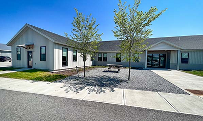 Single-story office building with ell addition, two entrances, with concrete sidewalks, young trees and picnic table out front. 