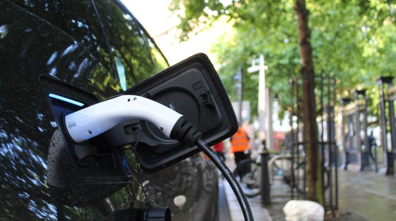 Close up of an electric vehicle charging port with charging cable attached. The car is on a city street with a sidewalk and trees in the background.